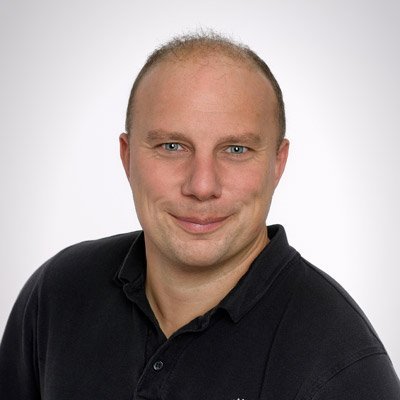 Ein lächelnder Mann mit kurzen Haaren trägt ein schwarzes Polohemd und blickt direkt in die Kamera.