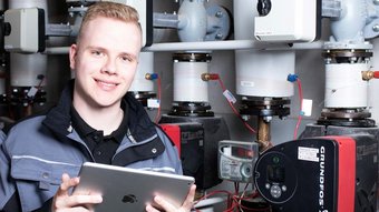 Ein junger Anlagenmechaniker vor einer Heizungsanlage, der ein Tablet hält 