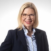 Eine lächelnde Frau mit blonden Haaren trägt eine Brille und einen dunklen Blazer, vor einem hellen Hintergrund.