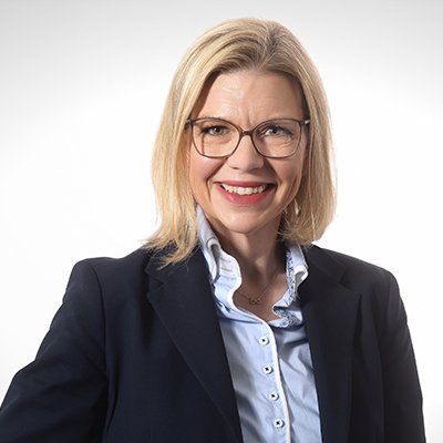 Eine lächelnde Frau mit blonden Haaren trägt eine Brille und einen dunklen Blazer, vor einem hellen Hintergrund.