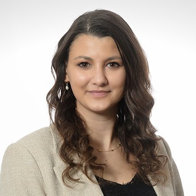 Eine junge Frau mit lockigem Haar trägt eine helle Jacke und lächelt freundlich in die Kamera.