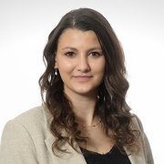 Eine junge Frau mit lockigem Haar trägt eine helle Jacke und lächelt freundlich in die Kamera.