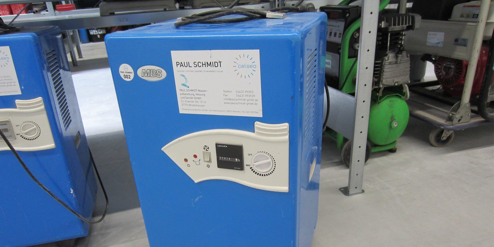 Ein blauer Heizungsgerät mit einem Bedienfeld und einem Aufkleber von Paul Schmidt steht in einer Werkstatt.