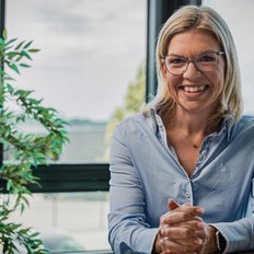 In einem modernen Büro lächelt eine Frau in einem hellblauen Hemd, während sie ihre Hände auf dem Tisch faltet.