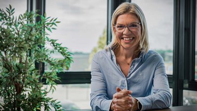 In einem modernen Büro lächelt eine Frau in einem hellblauen Hemd, während sie ihre Hände auf dem Tisch faltet.