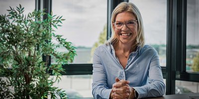 In einem modernen Büro lächelt eine Frau in einem hellblauen Hemd, während sie ihre Hände auf dem Tisch faltet.