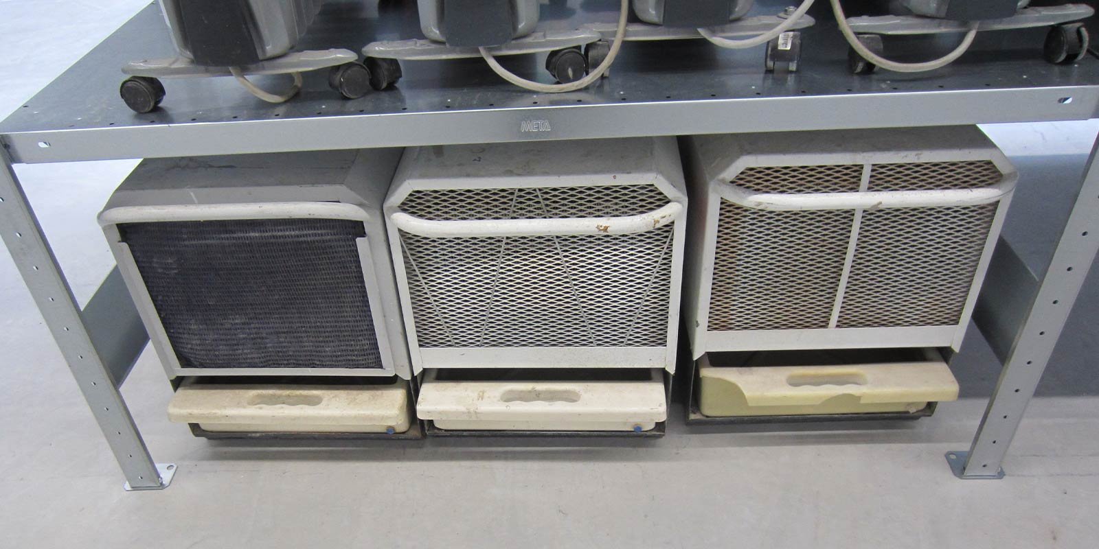 Ein Metallregal trägt drei Geräte mit Gitterstrukturen, die in verschiedenen Zuständen der Abnutzung sind.