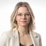 Eine junge Frau mit blonden, lockigen Haaren und Brille trägt einen eleganten Blazer und schaut direkt in die Kamera.