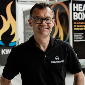 Ein lächelnder Mann in einem schwarzen Poloshirt steht vor einem Heizungsanhänger mit dem Schriftzug 'HEAT BOX'.