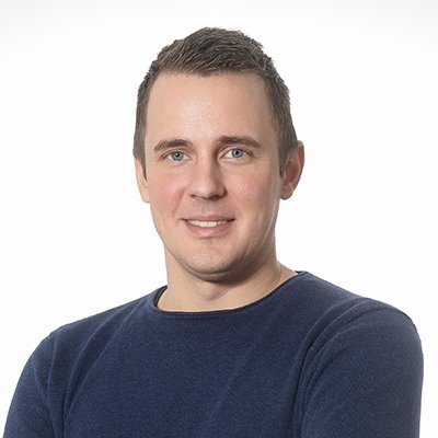 Ein junger Mann mit kurzem, braunem Haar lächelt freundlich in die Kamera, während er einen blauen Pullover trägt.
