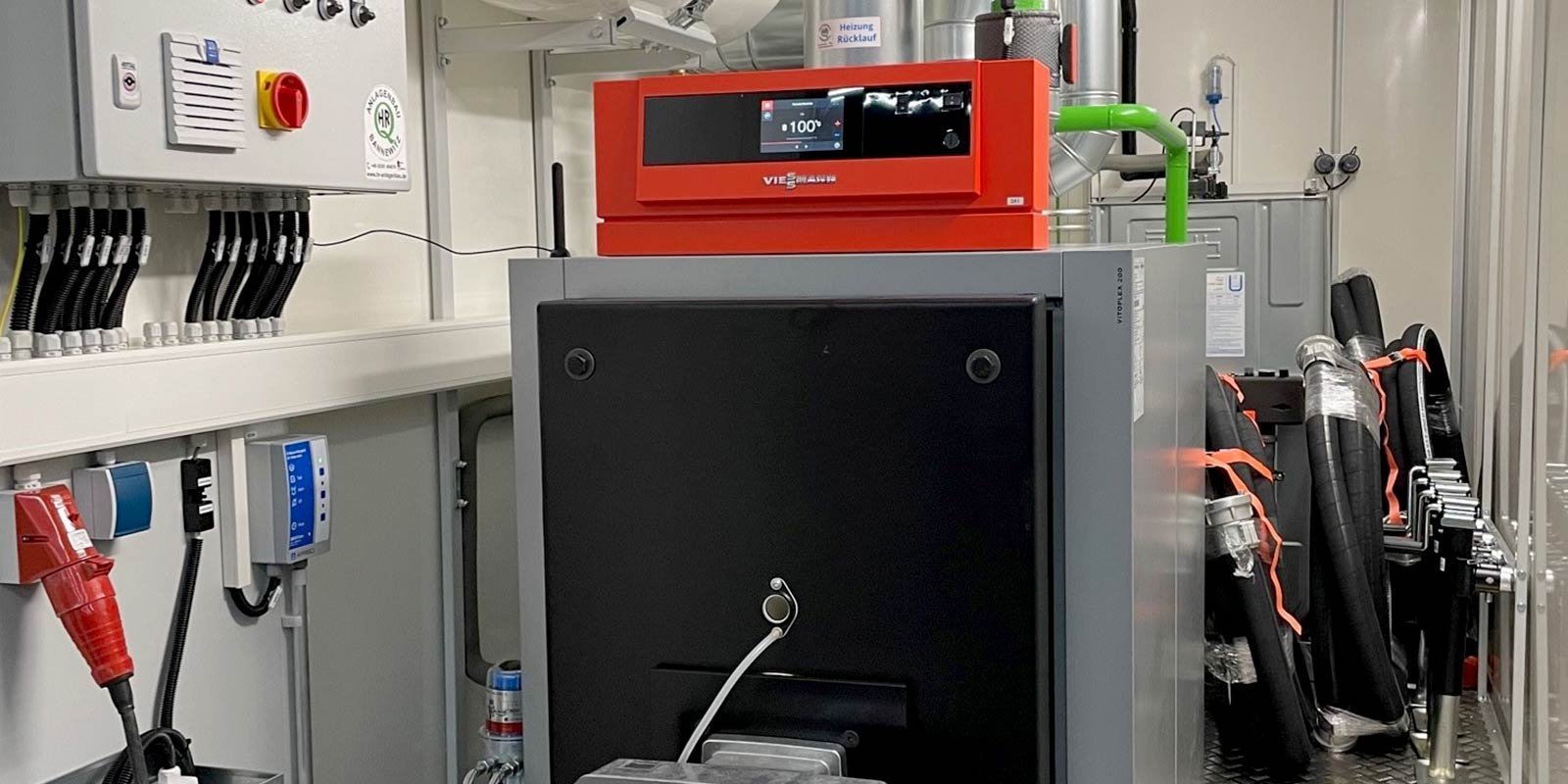 Ein rotes Steuergerät mit digitalem Display steht in einem technischen Raum, umgeben von verschiedenen Rohren und Kabeln.