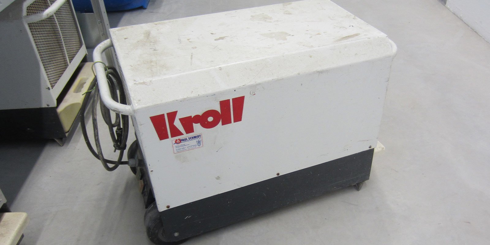 Ein weißer Kroll-Maschinenblock steht auf einem grauen Boden, umgeben von anderen Geräten in einem industriellen Raum.