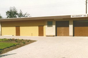 Ein modernes, gelbes Gebäude mit braunen Garagentoren und einem Schild mit der Aufschrift 'SCHMIDT KG' im Vordergrund.