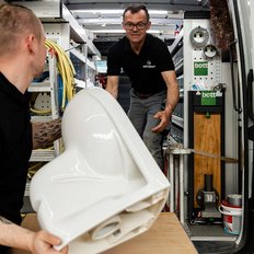 Im Inneren eines Lieferwagens stehen zwei Männer, einer trägt ein großes, weißes Teil, der andere hilft beim Anheben.