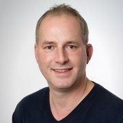 Ein lächelnder Mann mit kurzen, grauen Haaren trägt einen dunklen Pullover und posiert vor einem hellen Hintergrund.