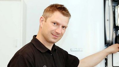 Ein Handwerker in schwarzem Shirt steht neben einer Heizungsanlage und hält ein Tablet in der Hand, während er lächelt.