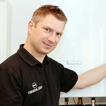 Techniker bei der Wartung einer Heizungsanlage im Einsatz Ein Handwerker in schwarzem Shirt steht neben einer Heizungsanlage und hält ein Tablet in der Hand, während er lächelt.