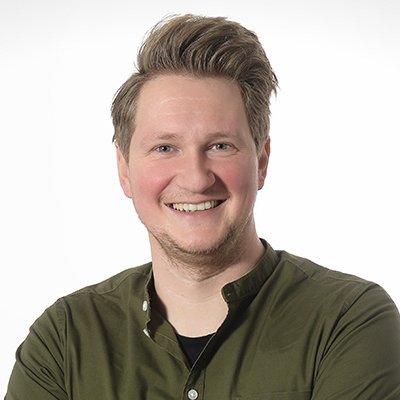 Ein lächelnder Mann mit blonden Haaren trägt ein grünes Hemd und steht vor einem hellen Hintergrund.