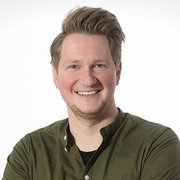Ein lächelnder Mann mit blonden Haaren trägt ein grünes Hemd und steht vor einem hellen Hintergrund.