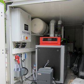 Ein technischer Raum mit einem großen Gerät, das mit Rohren und Kabeln verbunden ist, zeigt moderne Maschinen und Steuerungselemente.