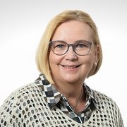 Eine lächelnde Frau mit blonden Haaren trägt eine Brille und ein gemustertes Hemd, vor einem hellen Hintergrund.