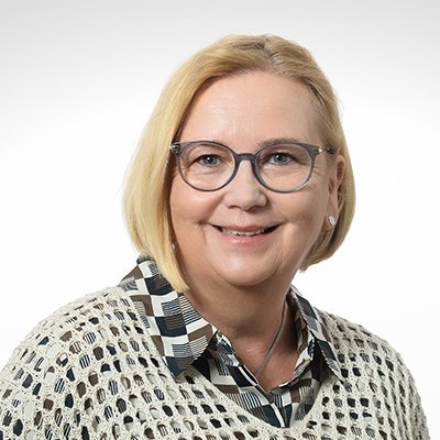 Eine lächelnde Frau mit blonden Haaren trägt eine Brille und ein gemustertes Hemd, vor einem hellen Hintergrund.