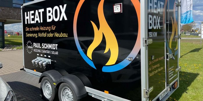 Ein schwarzer Heizungsanhänger mit dem Schriftzug 'HEAT BOX' und einem Flammenlogo, steht auf einem gepflasterten Parkplatz.