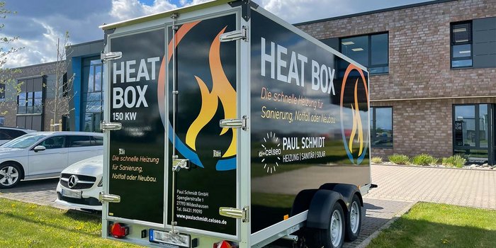Ein mobiler Heizcontainer mit der Aufschrift 'HEAT BOX' steht vor einem modernen Gebäude, umgeben von Autos und frischem Gras.