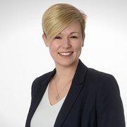 Eine lächelnde Frau mit blonden, kurzen Haaren trägt einen dunklen Blazer und eine helle Bluse vor einem neutralen Hintergrund.