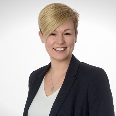 Eine lächelnde Frau mit blonden, kurzen Haaren trägt einen dunklen Blazer und eine helle Bluse vor einem neutralen Hintergrund.