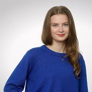 Eine junge Frau mit langen, lockigen Haaren lächelt freundlich in die Kamera, während sie einen blauen Pullover trägt.
