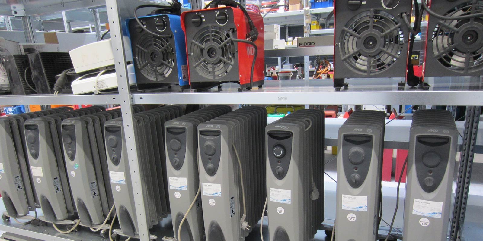 Eine Reihe von Heizgeräten steht ordentlich auf einem Regal, während einige Ventilatoren darüber angeordnet sind.