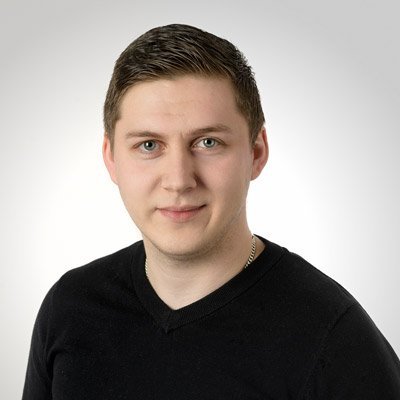 Ein junger Mann mit kurzem, dunklem Haar und einem freundlichen Lächeln trägt ein schwarzes Oberteil vor einem hellen Hintergrund.