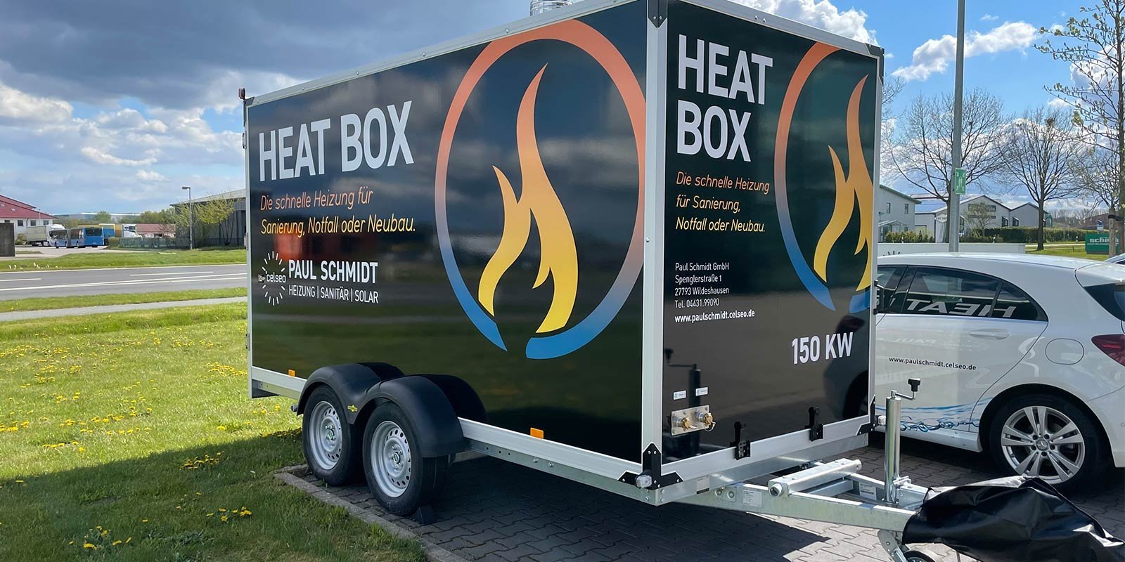 Ein Heizungsanhänger mit der Aufschrift 'HEAT BOX' steht auf einem Parkplatz, umgeben von grünem Gras und Bäumen.