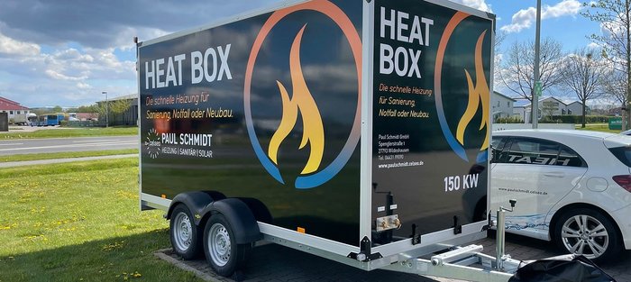 Ein Heizungsanhänger mit der Aufschrift 'HEAT BOX' steht auf einem Parkplatz, umgeben von grünem Gras und Bäumen.