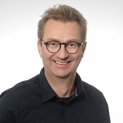 Ein lächelnder Mann mit Brille trägt ein schwarzes Hemd und steht vor einem hellen Hintergrund.