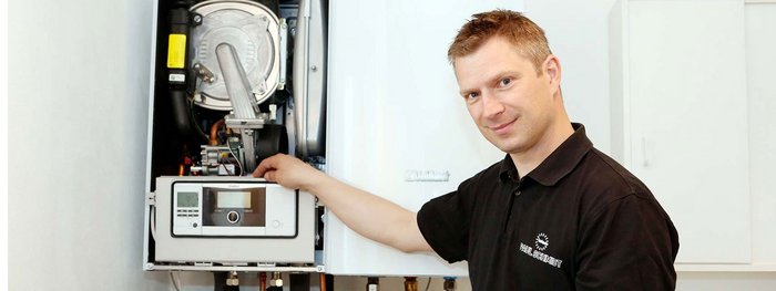 Ein Techniker steht vor einer Heizungsanlage und zeigt auf das Bedienfeld, während er freundlich lächelt.