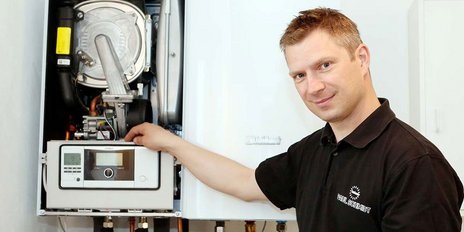 Ein Techniker steht vor einer Heizungsanlage und zeigt auf das Bedienfeld, während er freundlich lächelt.