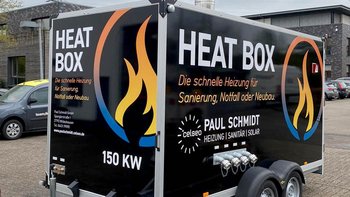 Ein Heizungsanhänger mit der Aufschrift 'HEAT BOX' steht auf einem Parkplatz, umgeben von mehreren Autos.